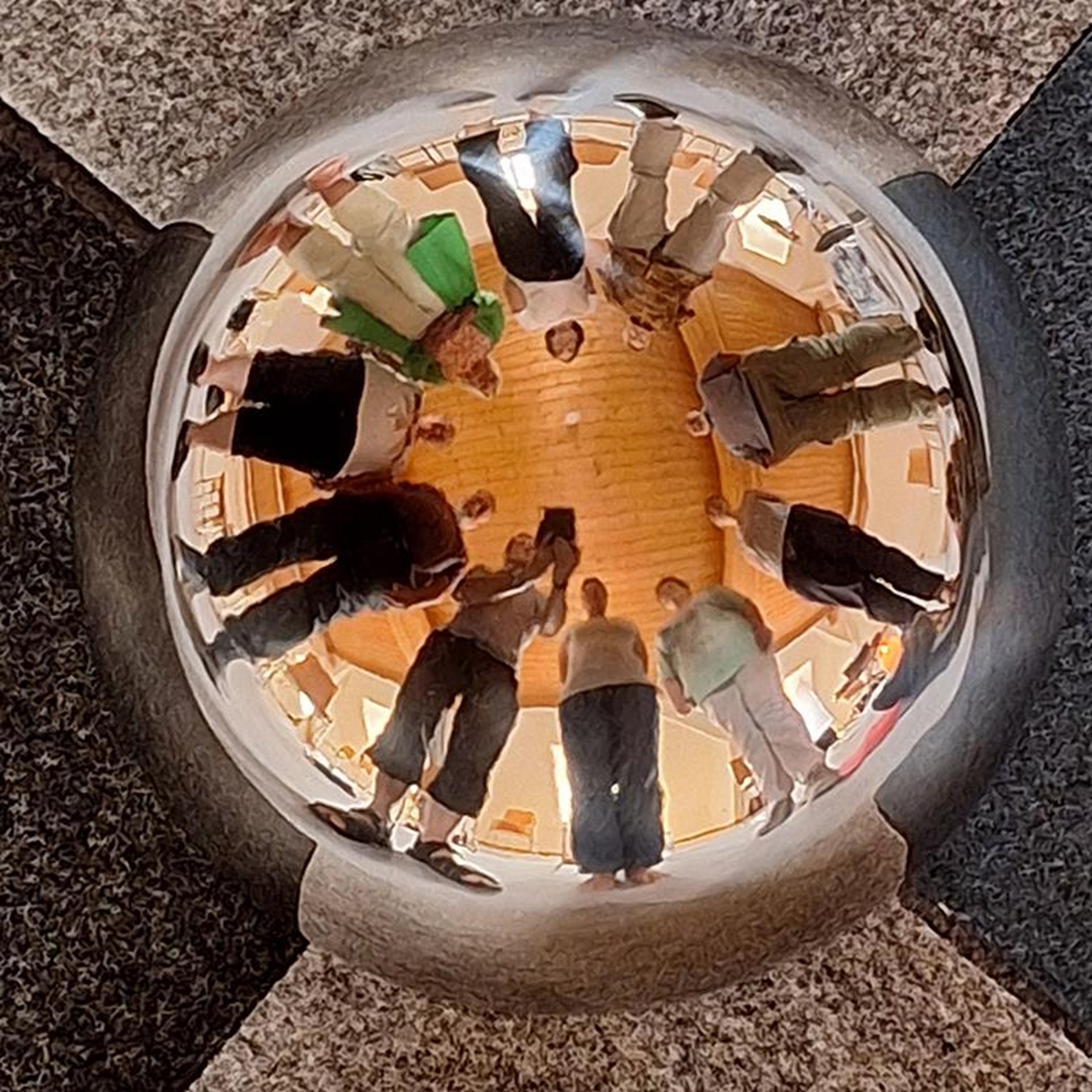 Eine Gruppe von Menschen spiegelt sich in einer polierten Metallkugel, die in einem geometrischen Steinboden eingebettet ist.