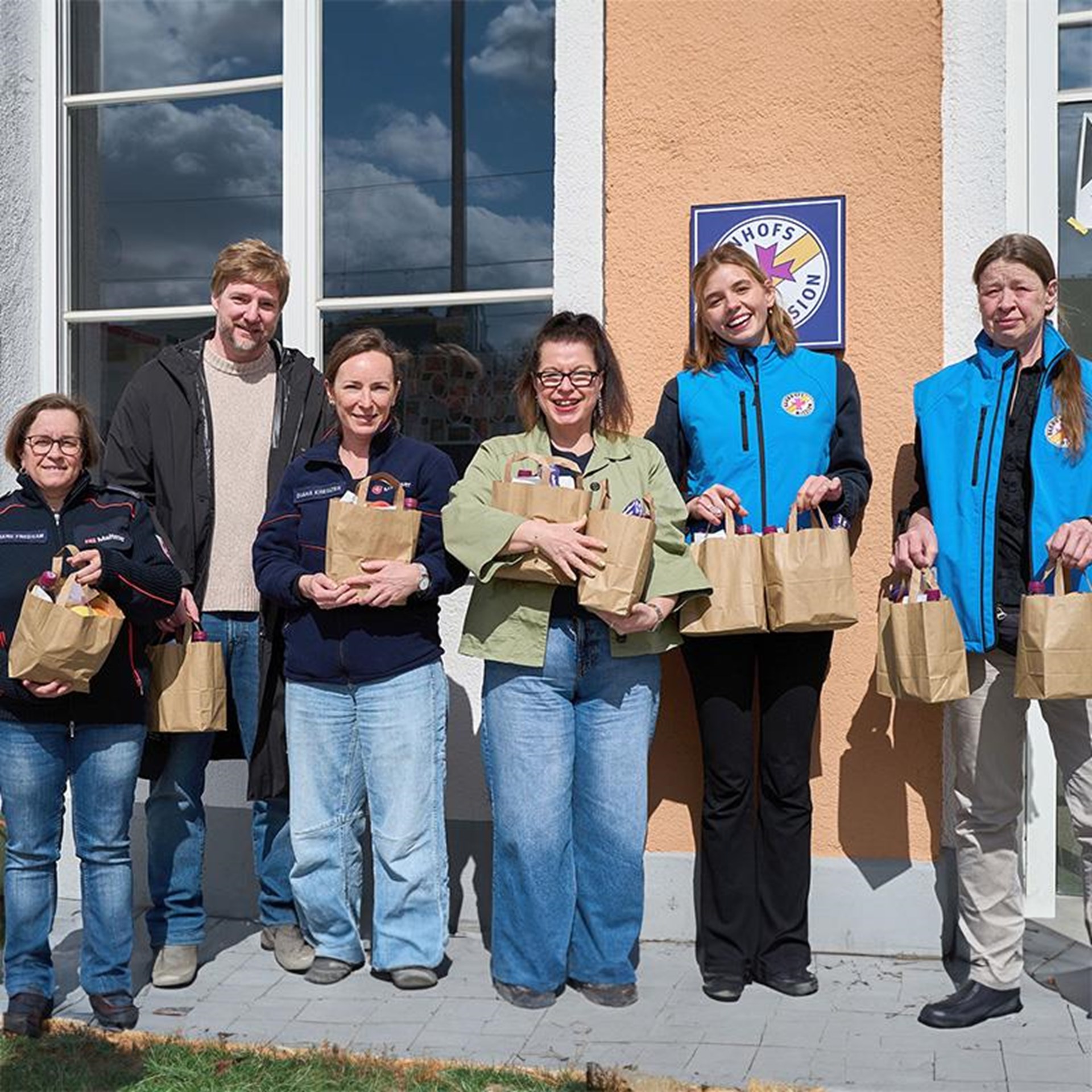 eine kleine Gruppe von Frauen und Männern stehen vor einer Bahnhofsmission und halten Papiertüten in der Hand in denen sich gespendete Lebensmittel befinden. 