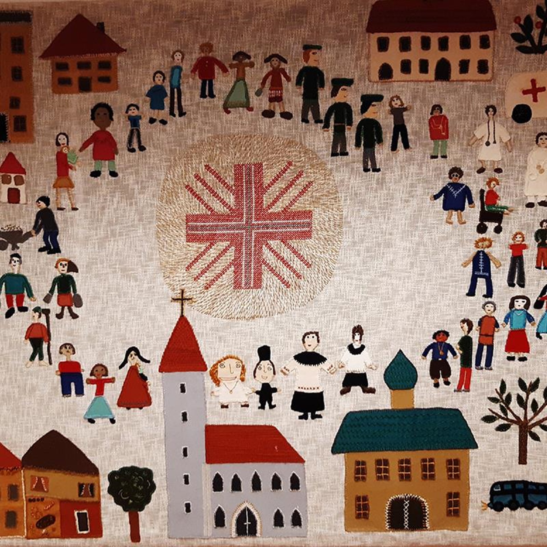 Eine kunstvolle Stickerei mit einer Dorfszene. Verschiedene Figuren, darunter Männer, Frauen und Kinder, sind zwischen bunten Häusern, Bäumen und Fahrzeugen dargestellt.