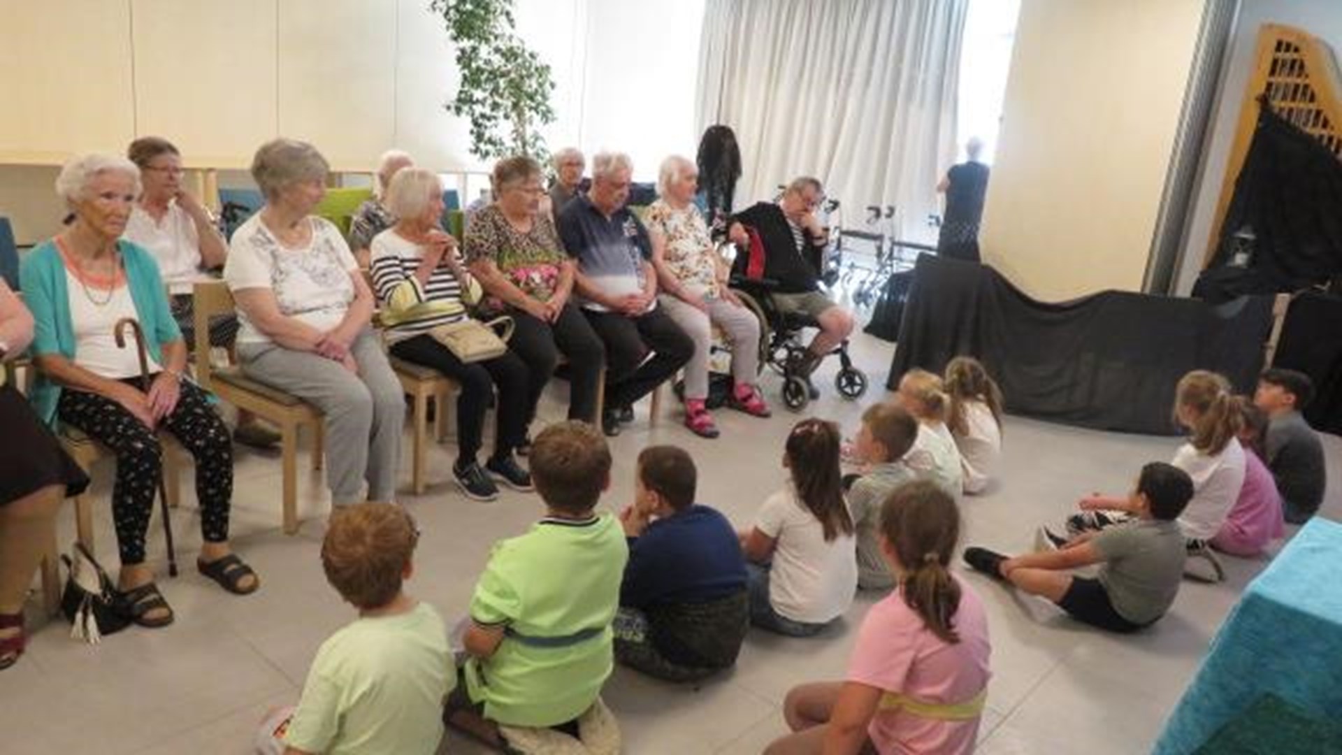Kinder sitzen im Gemeinschaftsraum des Caritas Seniorenheims St. Gisela vor den Bewohnenden auf dem Boden. Die Seniorinnen und Senioren verfolgen das Generationen-Theater zu den Bremer Stadtmusikanten von ihren Stühlen aus. Im Hintergrund ist eine kleine Spielfläche mit Tüchern vorbereitet.
