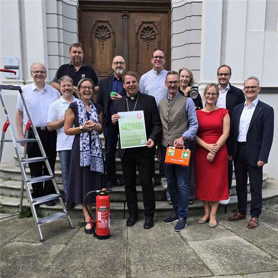 Gruppe von Mitarbeitenden vor einem historischen Eingang, darunter eine Person mit einem Plakat mit der Aufschrift „GESUNDHEITS-TAGE im Bistum Passau“ und „21. September bis 2. Oktober“. Die Gruppe präsentiert die Gesundheitstage bei der Caritas Passau mit Erste-Hilfe-Koffer und Feuerlöscher.