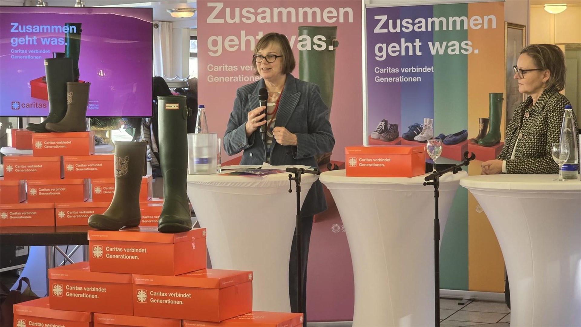 Zwei Personen stehen an Stehtischen mit Mikrofonen vor Bannern mit dem Text 'Zusammen geht was. Caritas verbindet Generationen.' und mehreren gestapelten orangen Schuhkartons mit Gummistiefeln darauf.