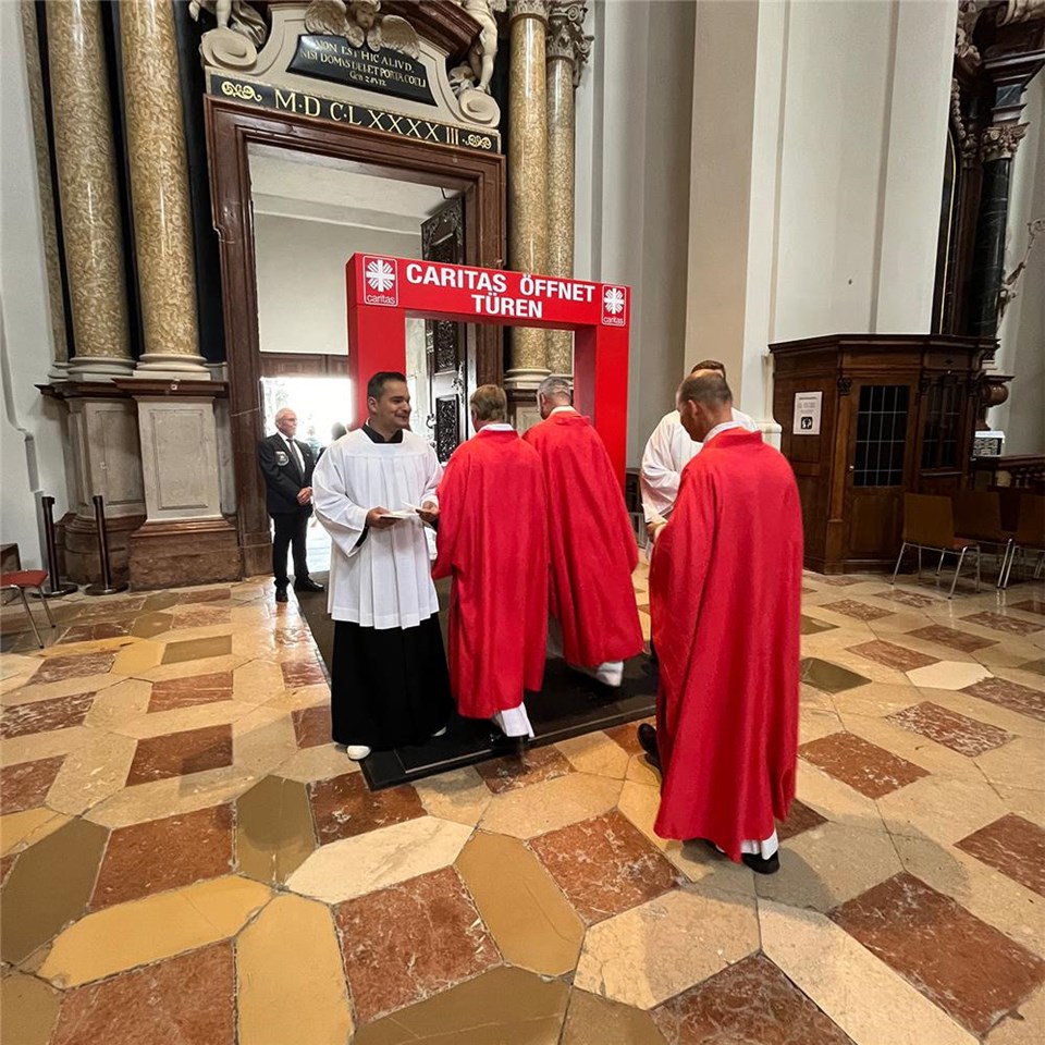 Drei Priester gehen in einem Dom durch ein rotes Tor mit der Aufschrift: "Caritas öffnet Türen".