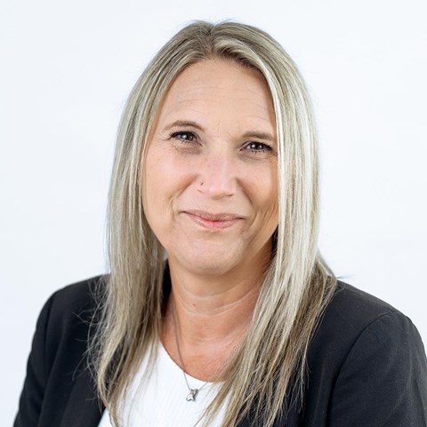 Eine Frau mit glatten, schulterlangen, blondierten Haaren lächelt dezent. Sie trägt einen schwarzen Blazer über einem weißen Oberteil und eine feine Halskette mit kleinem Anhänger vor einem weißen Hintergrund.