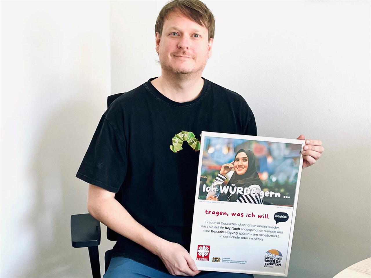 Ein Mann in schwarzem T-Shirt hält ein Plakat mit einer Kampagne gegen Diskriminierung von Frauen, die ein Kopftuch tragen.