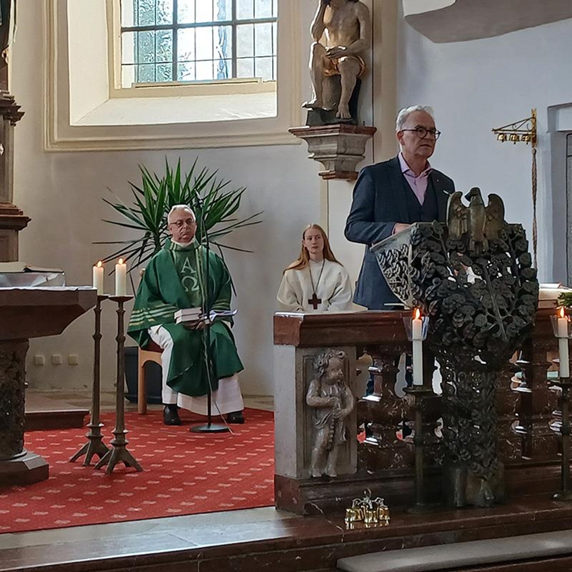 auf dem Foto sieht man den Altarbereich einer Kirche, im Hintergrund sitzt der Pfarrer auf einem Hocker, am Podium steht ein Mann. 