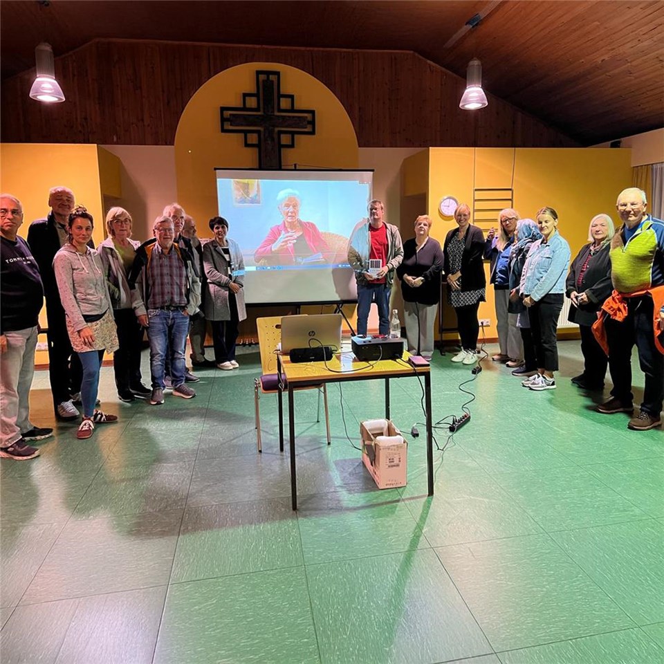 Eine Gruppe von Personen steht vor einer Leinwand. In der Mitte steht ein Tisch mit einem Beamer und Laptop.