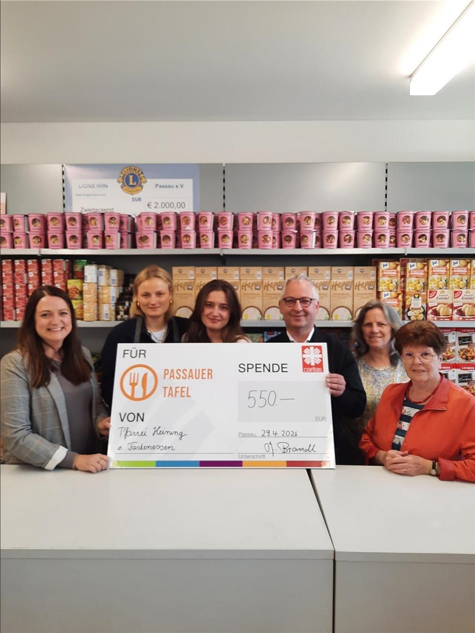 Sechs Personen stehen in den Räumen der Passauer Tafel vor Regalen mit Lebensmitteln. In der Mitte halten sie einen Spendenscheck der Pfarrei Heining über 550 Euro für die Passauer Tafel.