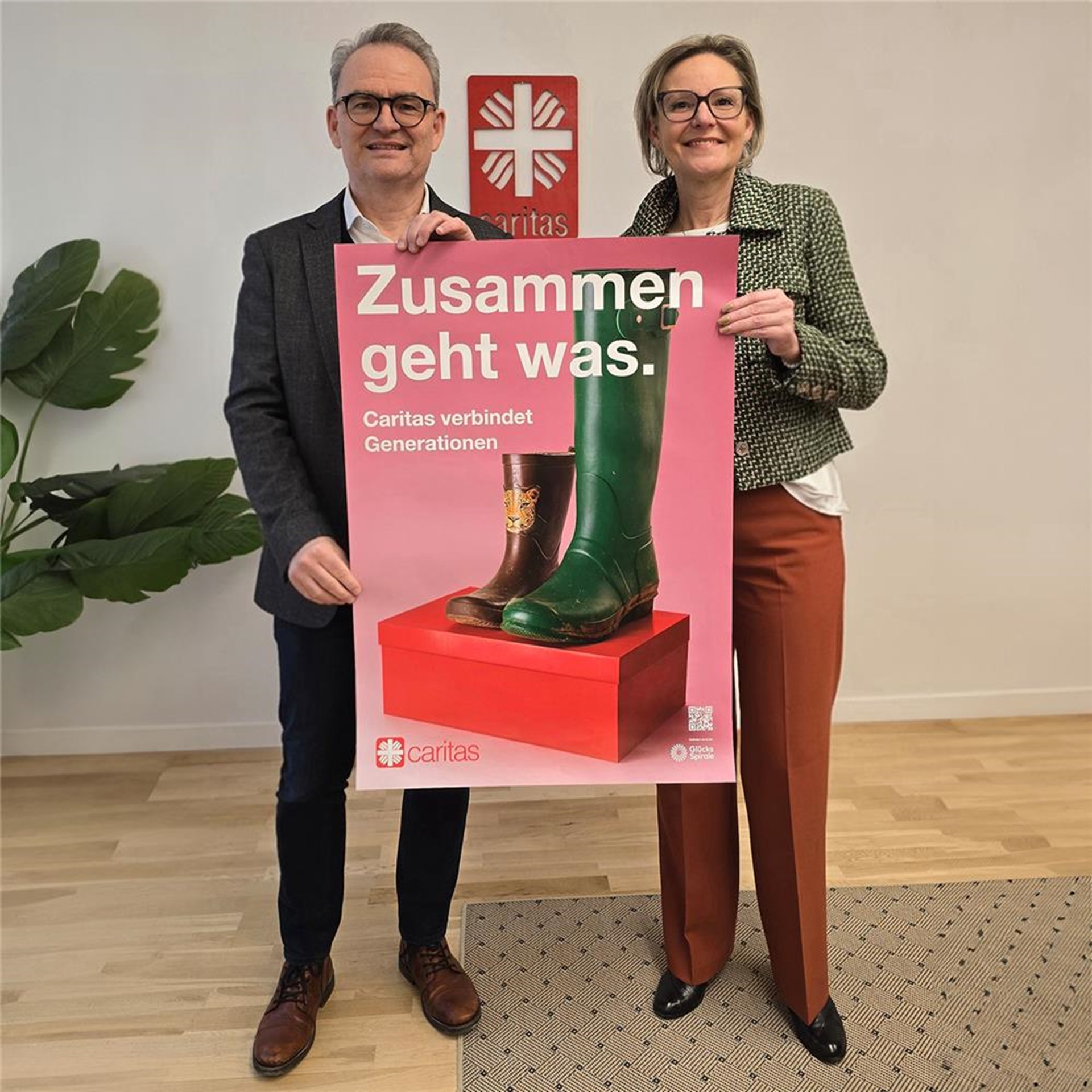Die Passauer Caritasvorstände Andrea Anderlik und Stefan Seidererstehen in einem hellen Raum und halten ein Plakat der Caritas-Jahreskampagne 2026 mit der Aufschrift „Zusammen geht was. Caritas verbindet Generationen“. Auf dem Plakat sind ein Kinderstiefel und ein Erwachsenengummistiefel auf einem roten Podest abgebildet, im Hintergrund ist das Caritas-Logo zu sehen.