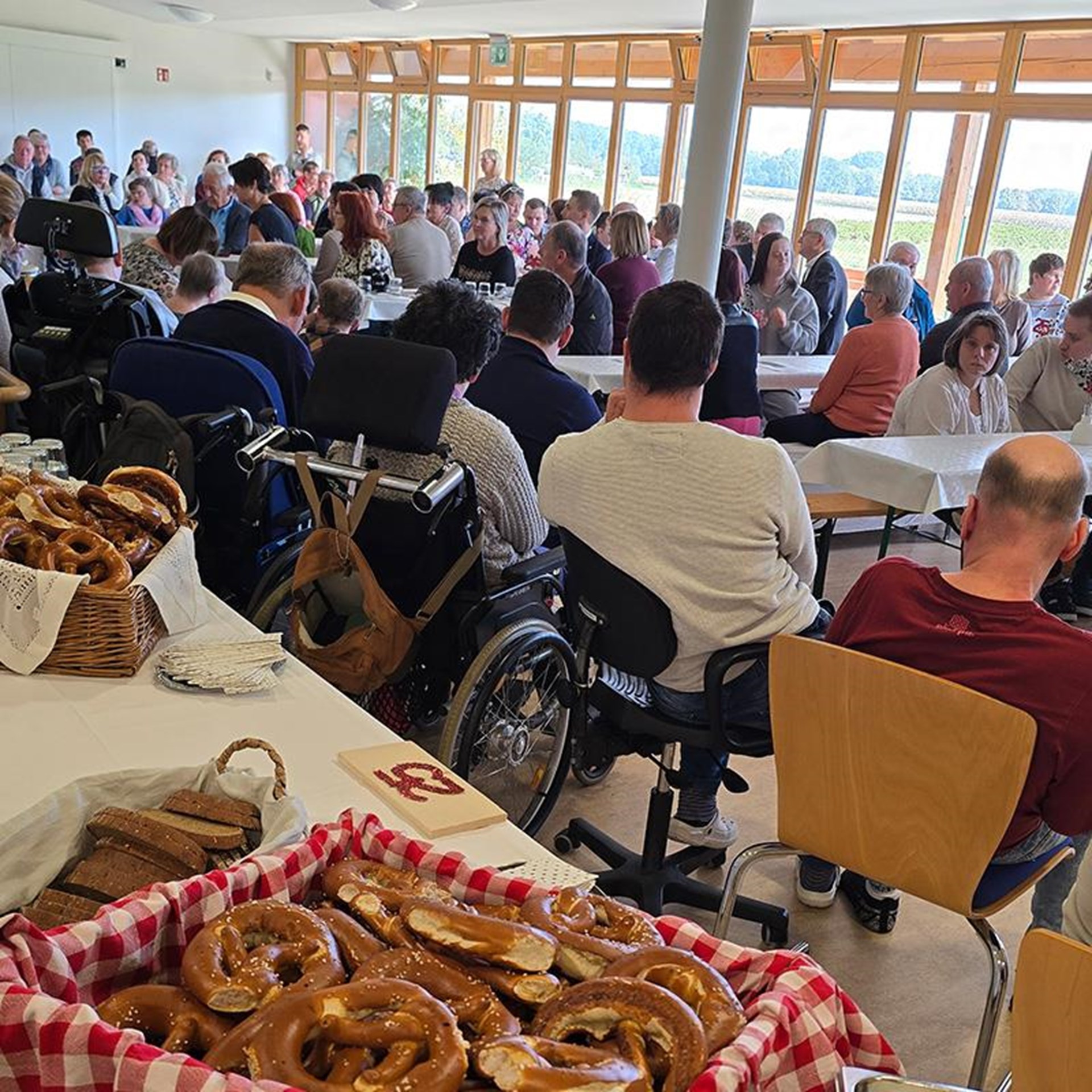 auf dem Bild ist ein großer Saal zu sehen. An Bierbänken und Tischen sitzen eine größere Anzahl von Personen. Einige davon sitzen im Rollstuhl. Auf der rechten Seite ist eine große Fensterfront. Auf der 