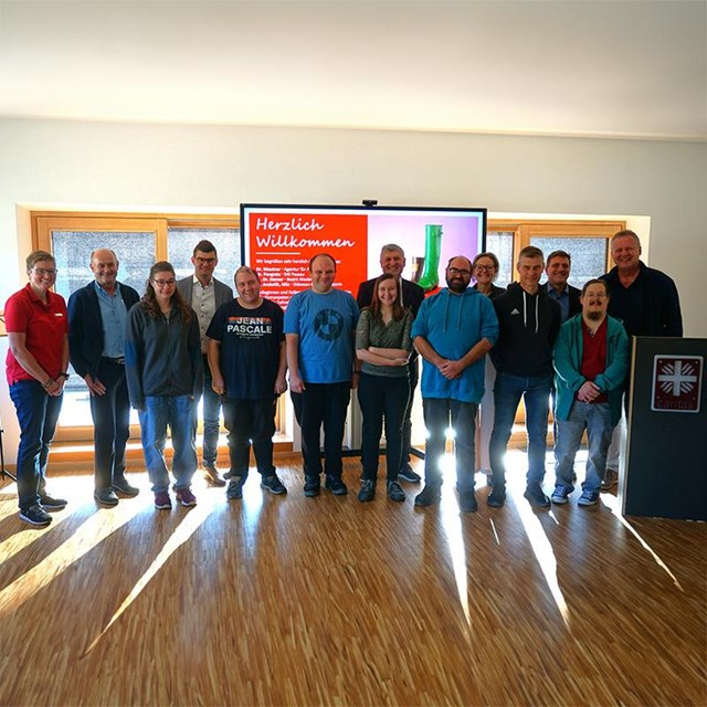 auf dem Bild ist eine Gruppe von 13 Frauen und Männern zu sehen, auf der rechten Seite steht ein Rednerpult mit dem Caritas Logo drauf. Im Hintergrund ist eine Leinwand auf der steht auf rotem Hintergrund Herzlich Willkommen.  