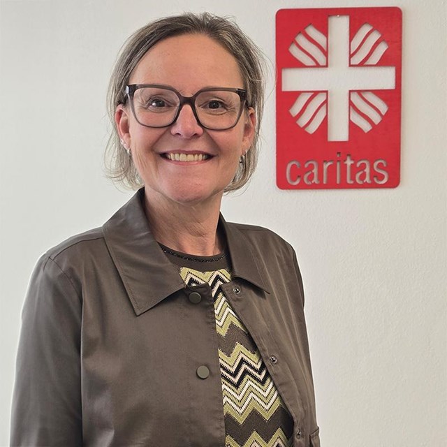 auf dem Bild ist eine Frau zu sehen mit kinnlangen Haaren. Sie lächelt in die Kamera, im Hintergrund ist eine weisse Wand und links davon das Caritaslogo
