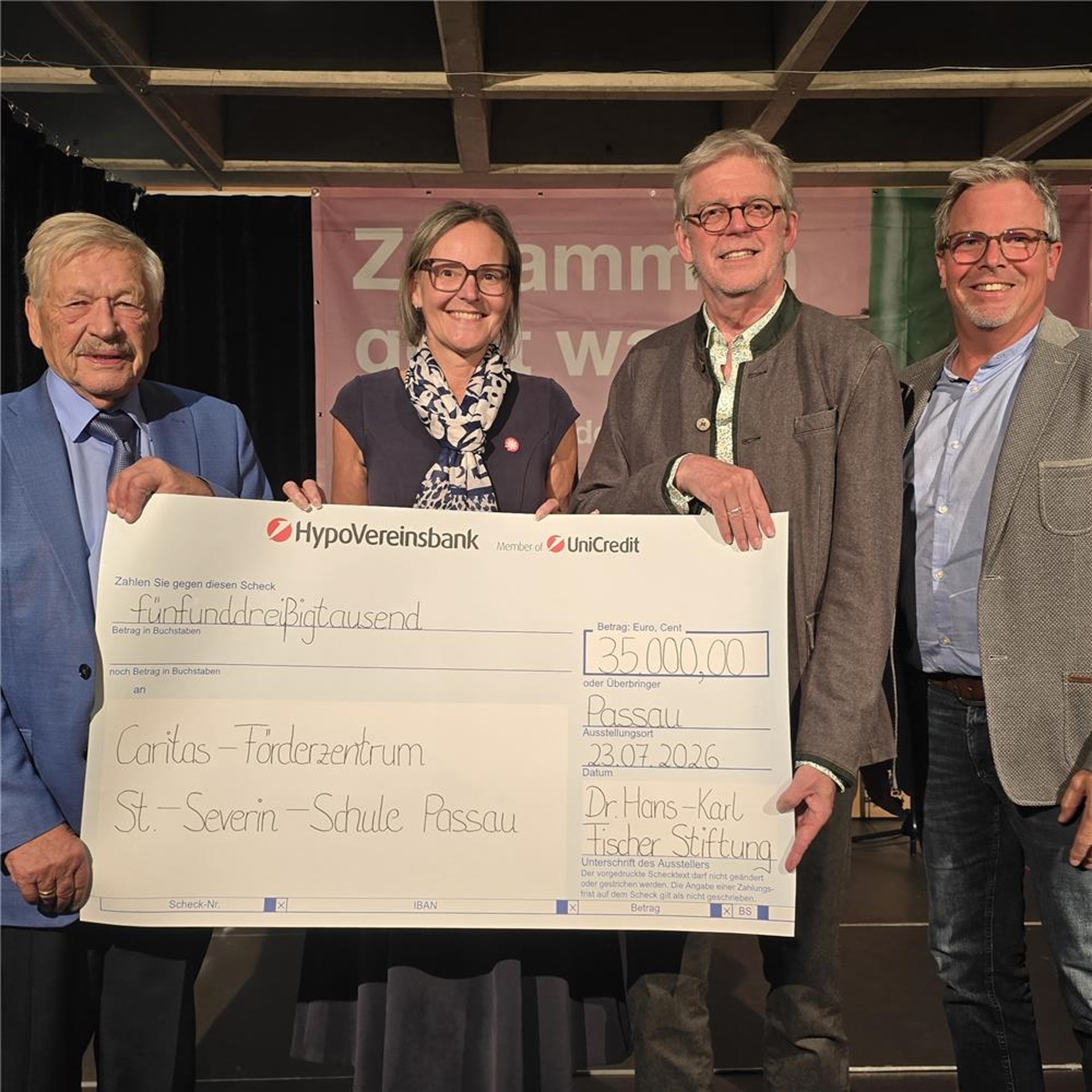 Vier Personen stehen auf einer Bühne und präsentieren einen großen Spendenscheck über 35.000 Euro der Dr. Hans-Karl Fischer Stiftung für einen neuen Schulbus des Caritas-Förderzentrums St. Severin.