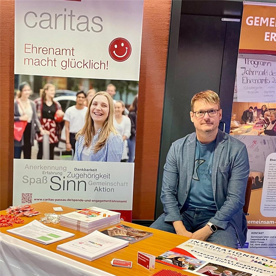 Eine Person sitzt bei einem Stand der Caritas. Am Stand liegen verschiedenste Informationsmaterialen wie Plakate usw. 