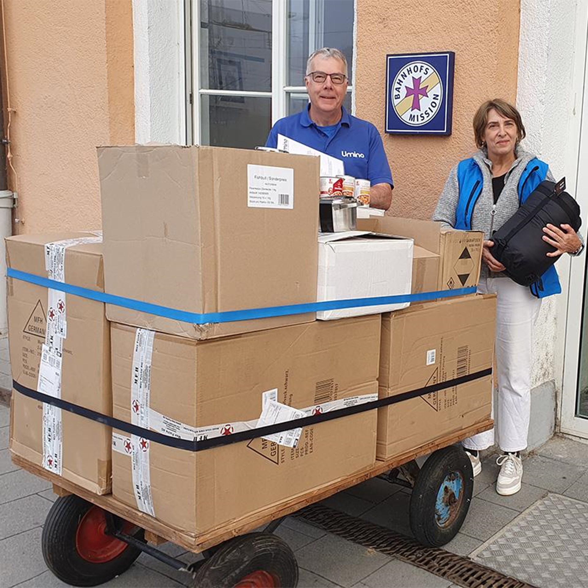 auf dem Bild ist ein Mann und eine Frau zu sehen, vor ihnen steht ein größerer Handwagen auf dem mehrere große Kartons sind. Der Mann hält einige Dosensuppen in der Hand, die Frau einen schwarzen Schlafsack. Im Hintergrund ist das lachsfarbene Gebäude der Bahnhofsmission zu sehen