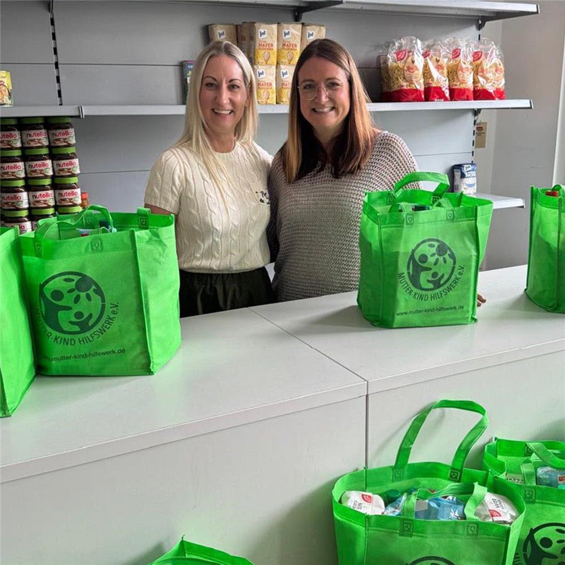 Zwei Personen stehen vor einem gefüllten Regal mit Lebensmittel. Vor ihnen stehen mehrere grüne Taschen die ebenfalls mit Lebensmitteln gefüllt sind.