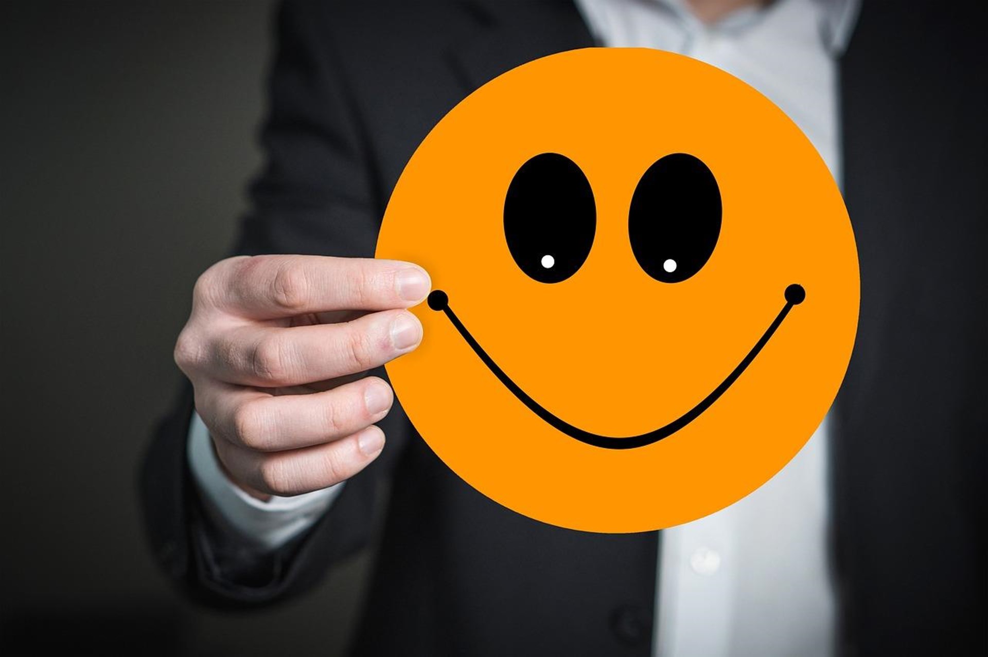 Eine Person im Anzug hält ein großes, orangefarbenes Smiley-Gesicht vor sich. Das Bild vermittelt ein Gefühl von Freundlichkeit und positiver Stimmung.