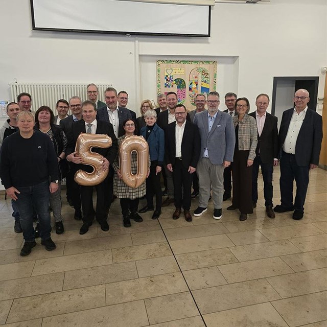 auf dem Bild steht eine Gruppe von Frauen und Männern, ein Mann und eine Frau hält jeweils die Zahl 5 und 0 in Form eines Luftballons in der Hand. Im Hintergrund ist ein buntes Bild mit der Zahl 50. rechts im Bild ist ein Schrank zu sehen und links im Hintergrund ein großer Heizkörper 