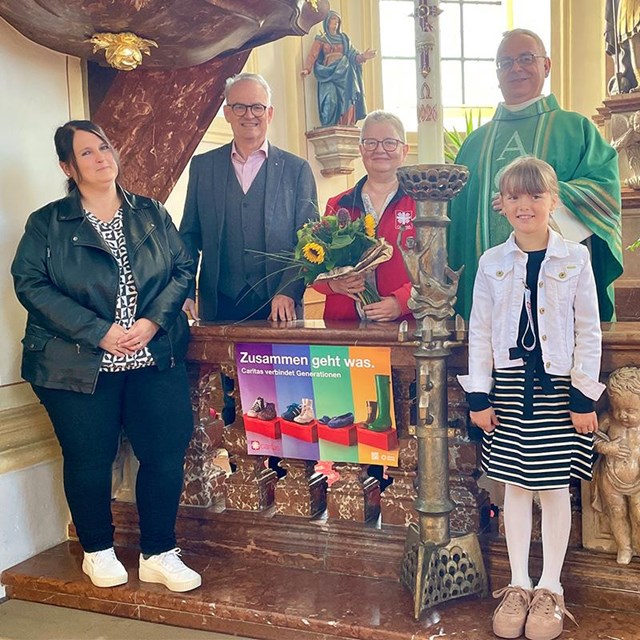 Fünf Personen – drei Frauen, ein Mann in Zivilkleidung und ein Priester in grünem Messgewand – stehen gemeinsam mit einem Mädchen vor dem Altar einer Kirche. Eine Frau in roter Jacke hält einen Blumenstrauß mit Sonnenblumen. Vor den Personen steht ein Plakat mit dem Caritas-Motto „Zusammen geht was. Caritas verbindet Generationen“. Im Hintergrund sind Kirchenfenster, Heiligenfiguren und Blumenschmuck zu sehen.