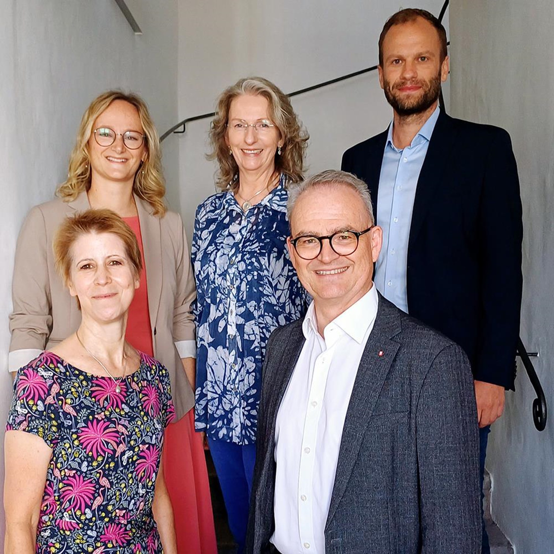 Fünf Personen stehen auf einer Treppe in einem hellen Gebäude und blicken freundlich in die Kamera. Zwei Männer im Anzug und drei Frauen in gemusterten Kleidern oder Blusen vermitteln einen professionellen, aber einladenden Eindruck – möglicherweise ein Team aus dem Gesundheits- oder Bildungsbereich.
