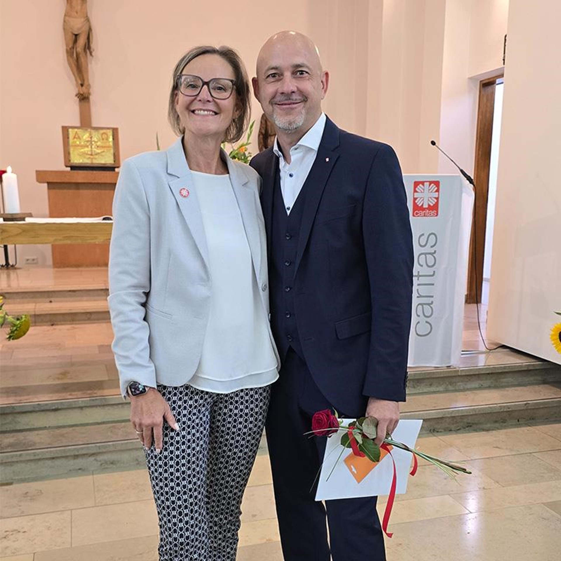 auf dem Bild ist eine Frau und ein Mann zu sehen welche in die Kamera lächeln, der Herr hält eine Mappe und eine Rose in der Hand