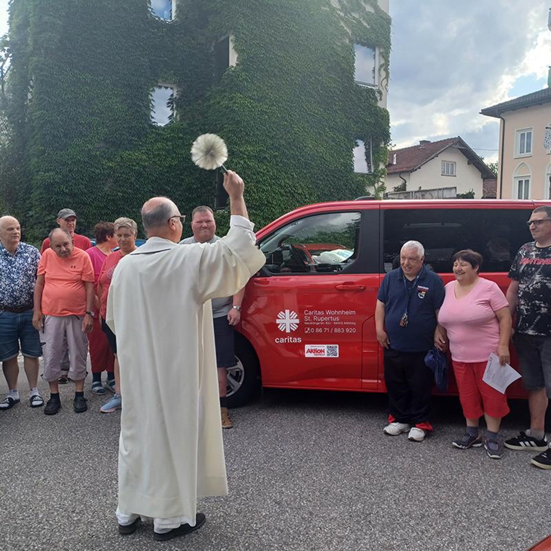 auf dem Bild ist ein Pfarrer zu sehen welcher einen roten Minibus segnet, links und rechts davon stehen Männer und Frauen