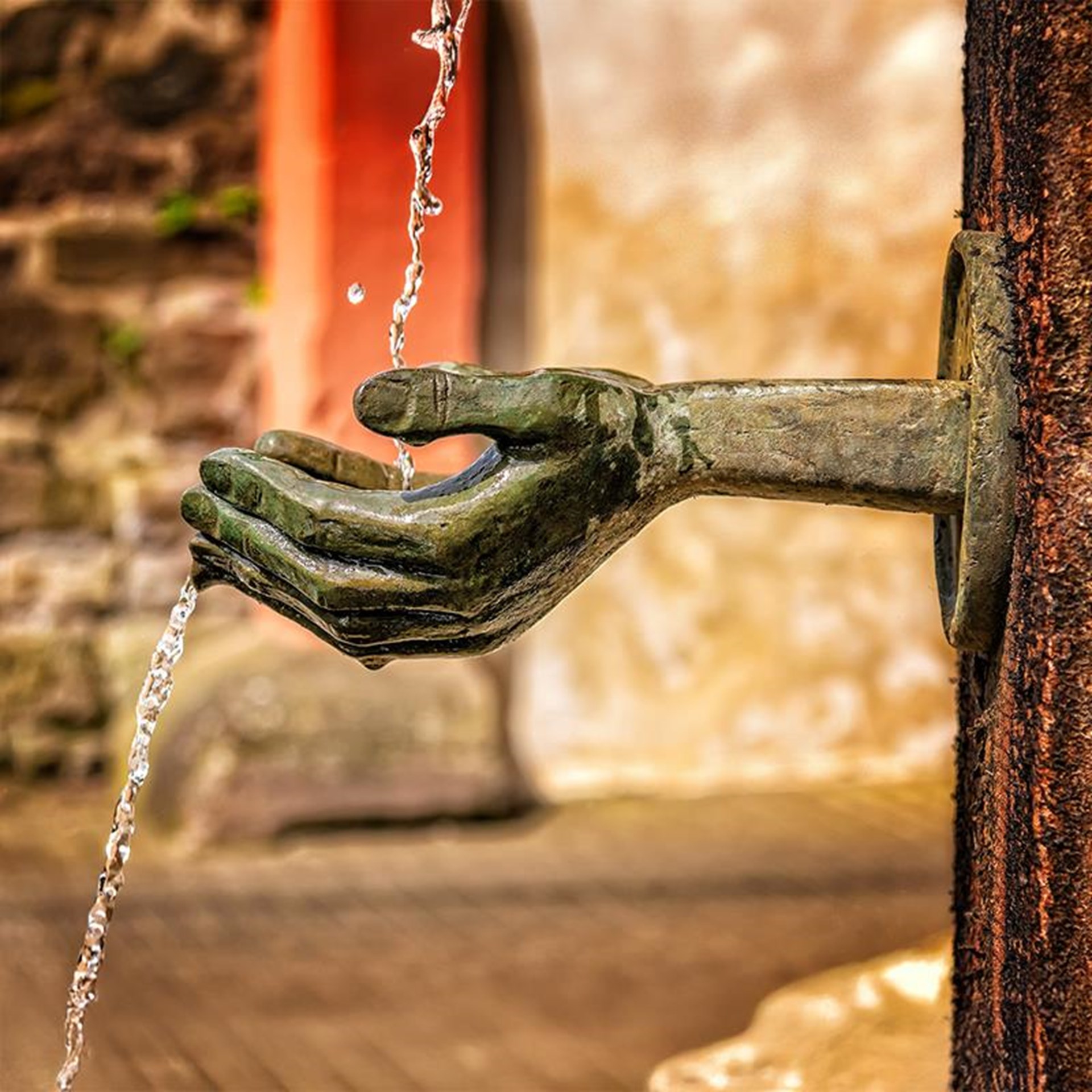 Ein kunstvoll gestalteter Brunnen mit einer bronzenen Hand, die aus einer Wand ragt und Wasser in ihre offene Handfläche auffängt, bevor es nach unten fließt. Die Szene spielt sich vor einer rustikale