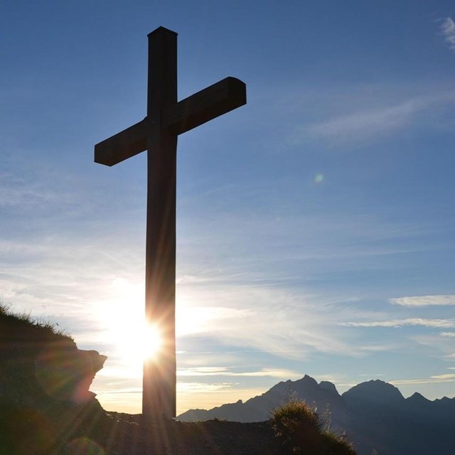 Ein hölzernes Gipfelkreuz steht silhouettenhaft vor blauem Himmel, während die Sonne direkt dahinter aufgeht oder untergeht. Im Hintergrund sind klare Berggipfel zu erkennen.