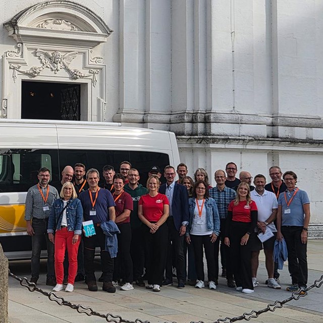 auf dem Bild ist eine Gruppe von Frauen und Männern zu sehen welche vor einem Minivan stehen. Im Hintergrund ist der Dom zu Passau 