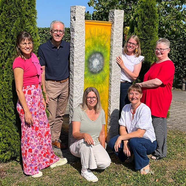 Auf einer sonnigen Grünfläche stehen und knien sechs Erwachsene vor einer farbenfrohen Stele aus zwei hohen Granitsäulen. Zwischen den Säulen hängt ein gelb-grünes Banner mit einem kreisförmigen Motiv in der Mitte. Vier Personen stehen links und rechts der Stele, zwei Personen knien davor. Alle blicken lächelnd in die Kamera. Die Gruppe trägt sommerliche Kleidung in verschiedenen Farben, darunter Rosa, Blau, Weiß und Rot. Im Hintergrund sind hohe Hecken, Bäume und ein wolkenloser blauer Himmel zu sehen