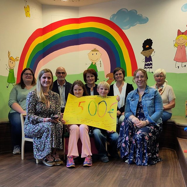 Sieben Erwachsene und zwei Kinder sitzen gemeinsam vor einer bunt bemalten Wand mit einem großen Regenbogen, einer Sonne und spielenden Kindern. Die beiden Kinder in der Mitte halten ein gelbes Plakat mit der Aufschrift „500 €“ und den Namen der Erstkommunionkinder aus Breitenberg-Sonnen in die Kamera. Das Foto zeigt die Spendenübergabe an das Ronald McDonald Haus in Passau.
