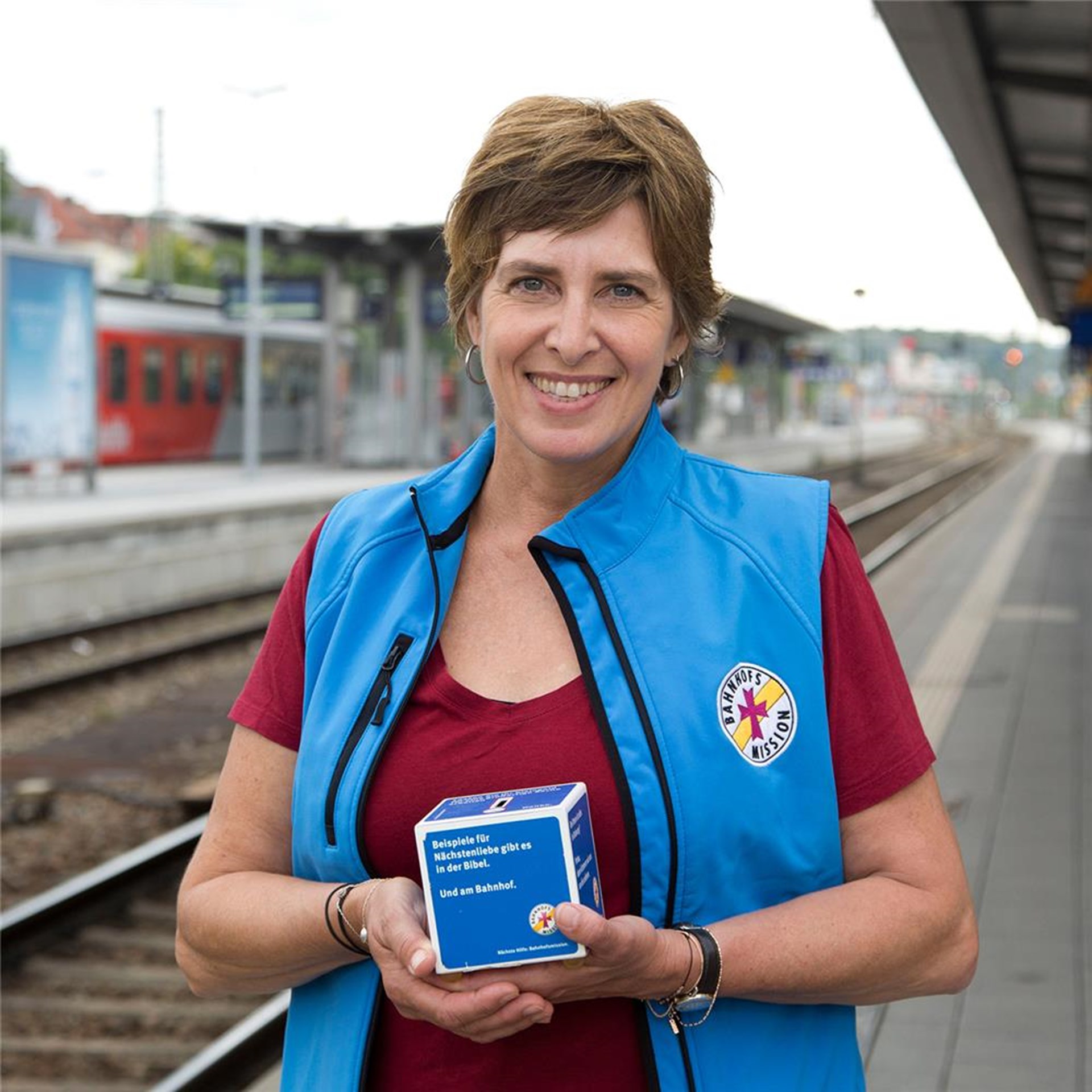 Eine Frau steht mit einem Würfel in der Hand am Bahnhof.