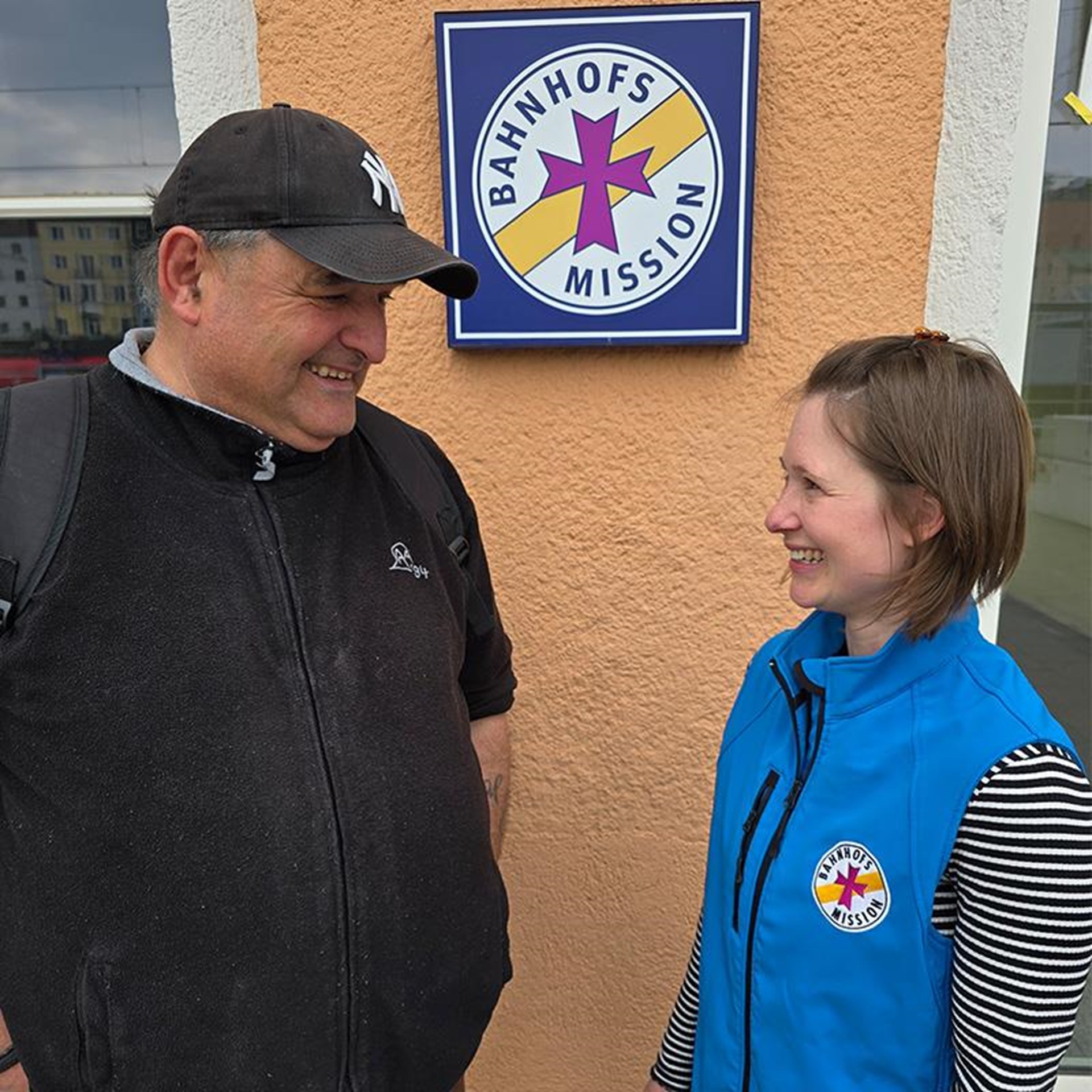 auf dem Bild ist ein Mann und eine Frau von der Bahnhofsmission zu sehen. Im Hintergrund das Bahnhofsgebäude