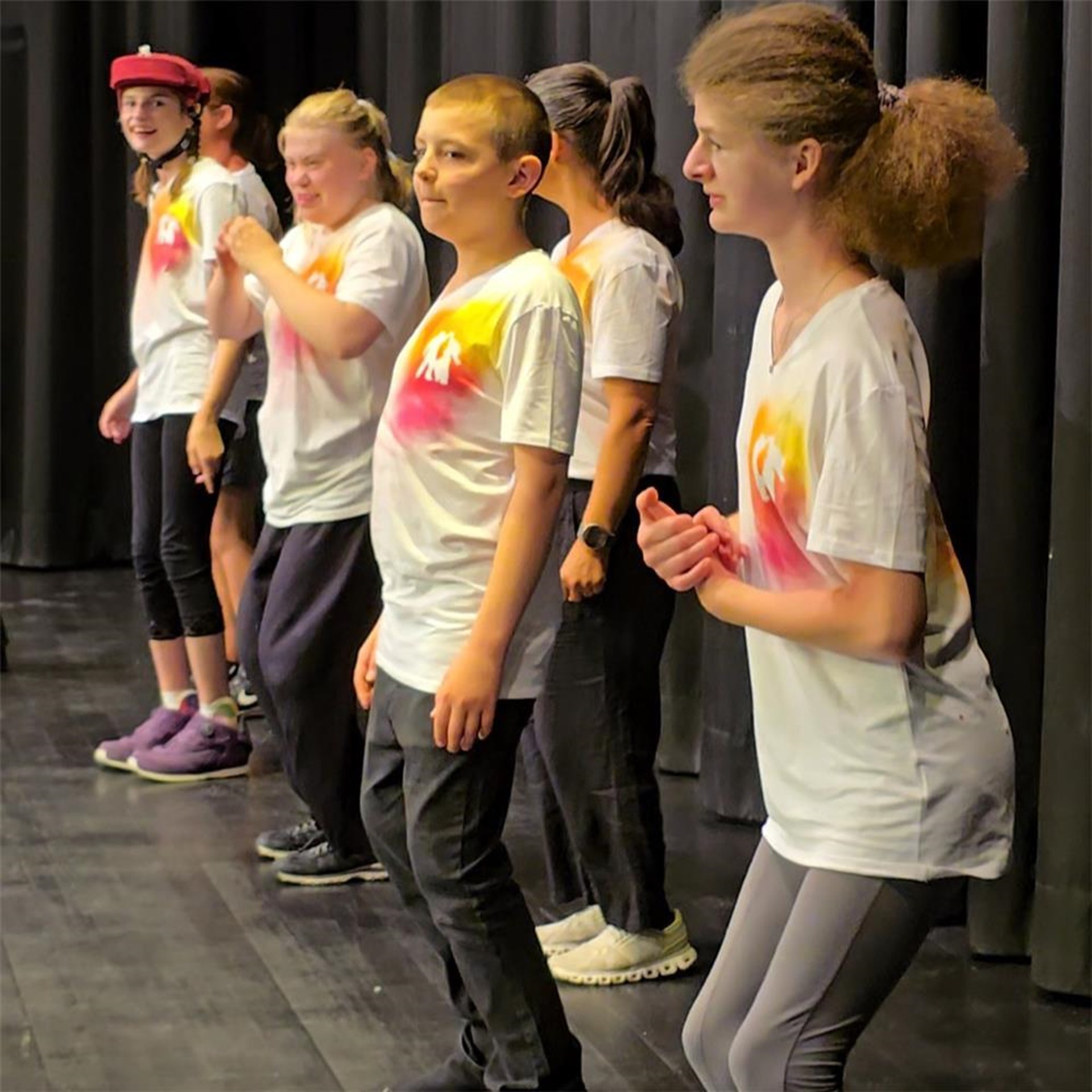 Eine Tanzgruppe der Caritas St. Ulrich-Schule Pocking steht auf der Bühne der Stadthalle Pocking. Die Schülerinnen und Schüler tragen helle T-Shirts mit farbigem Aufdruck und zeigen den Stehtanz „Ich bin ich“. Die Gruppe steht vor einem dunklen Bühnenvorhang und ist konzentriert bei ihrer Aufführung.