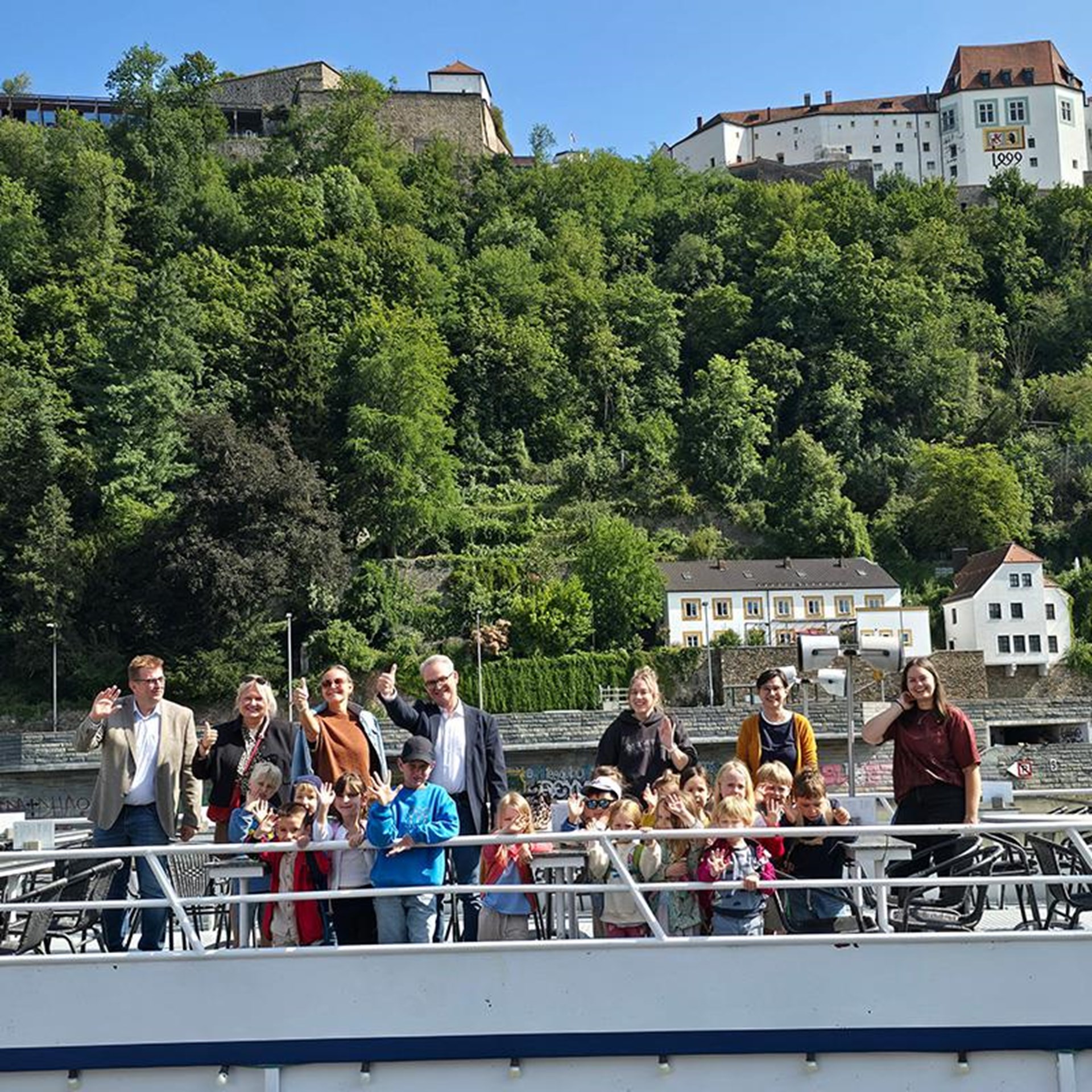 auf dem Bild ist ein Ausflugsschiff zu sehen von dem eine Gruppe Kindergartenkinder und 7 Erwachsene winken. Im Hintergrund ist oben auf einer Anhöhe eine Burg zu sehen