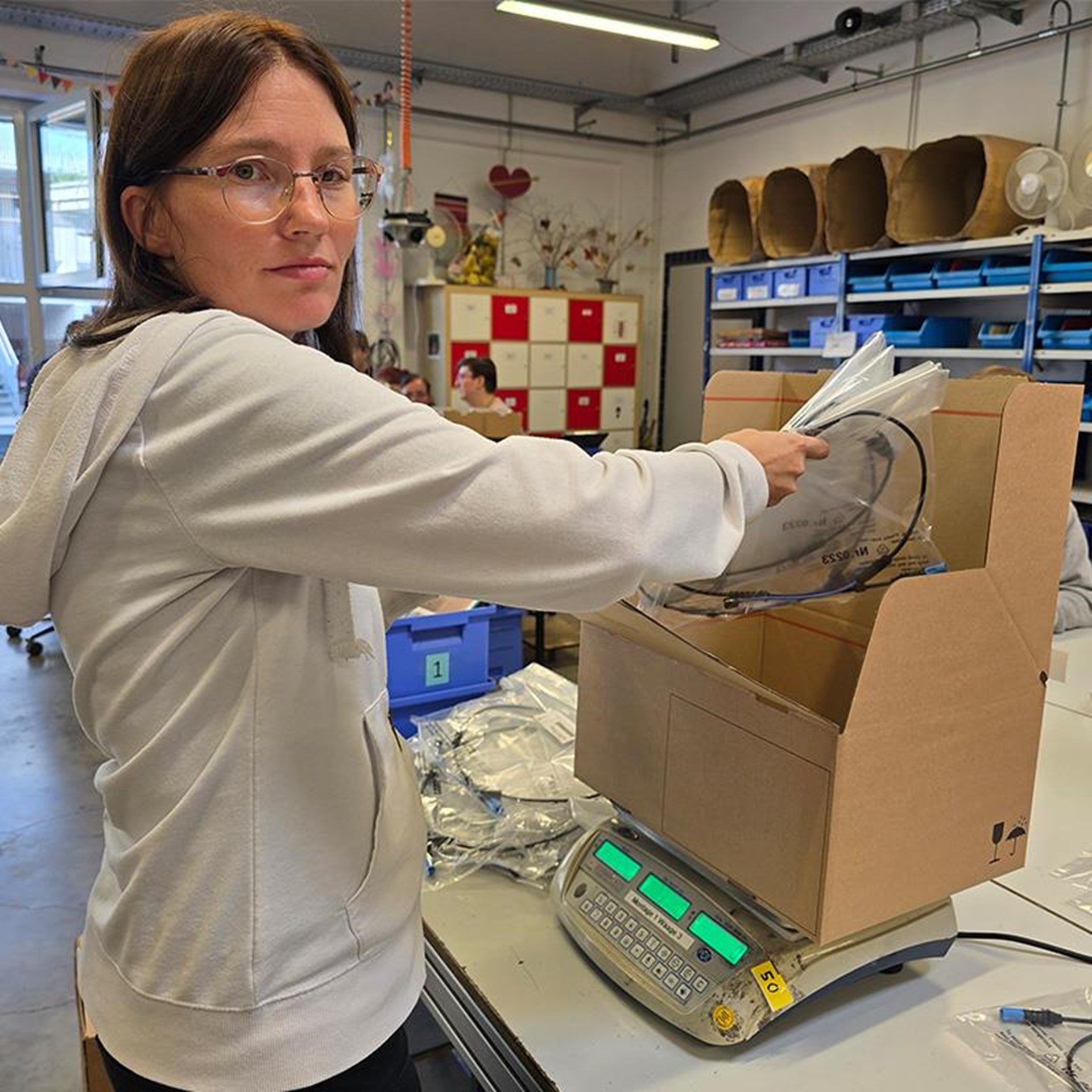 auf dem Bild ist eine junge Frau zu sehen in einer Werkstatt. Vor ihr steht auf dem Tisch eine Schachtel auf einer Waage welche sie mit Material befüllt. 