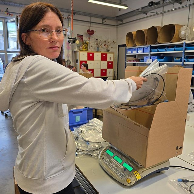 auf dem Bild ist eine junge Frau zu sehen in einer Werkstatt. Vor ihr steht auf dem Tisch eine Schachtel auf einer Waage welche sie mit Material befüllt. 