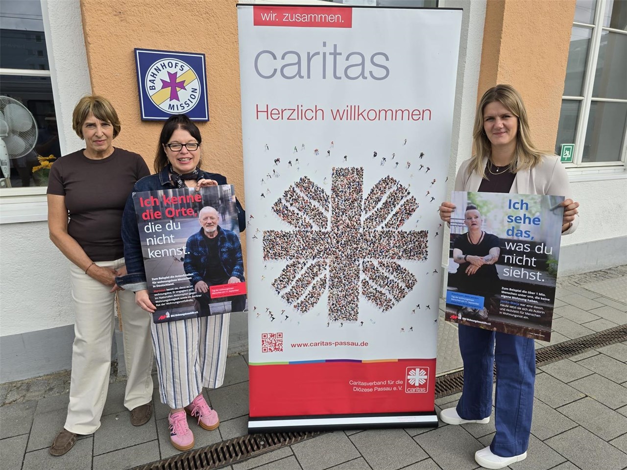 Drei Frauen stehen vor einem Caritas Aufsteller und halten Plakate zum Tag der Wohnungslosen in die Höhe.